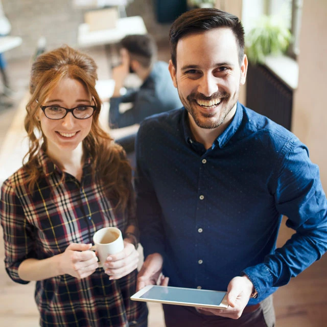 Eine weibliche Person mit Kaffeetasse und eine männliche Person mit Tablet, lächelnd